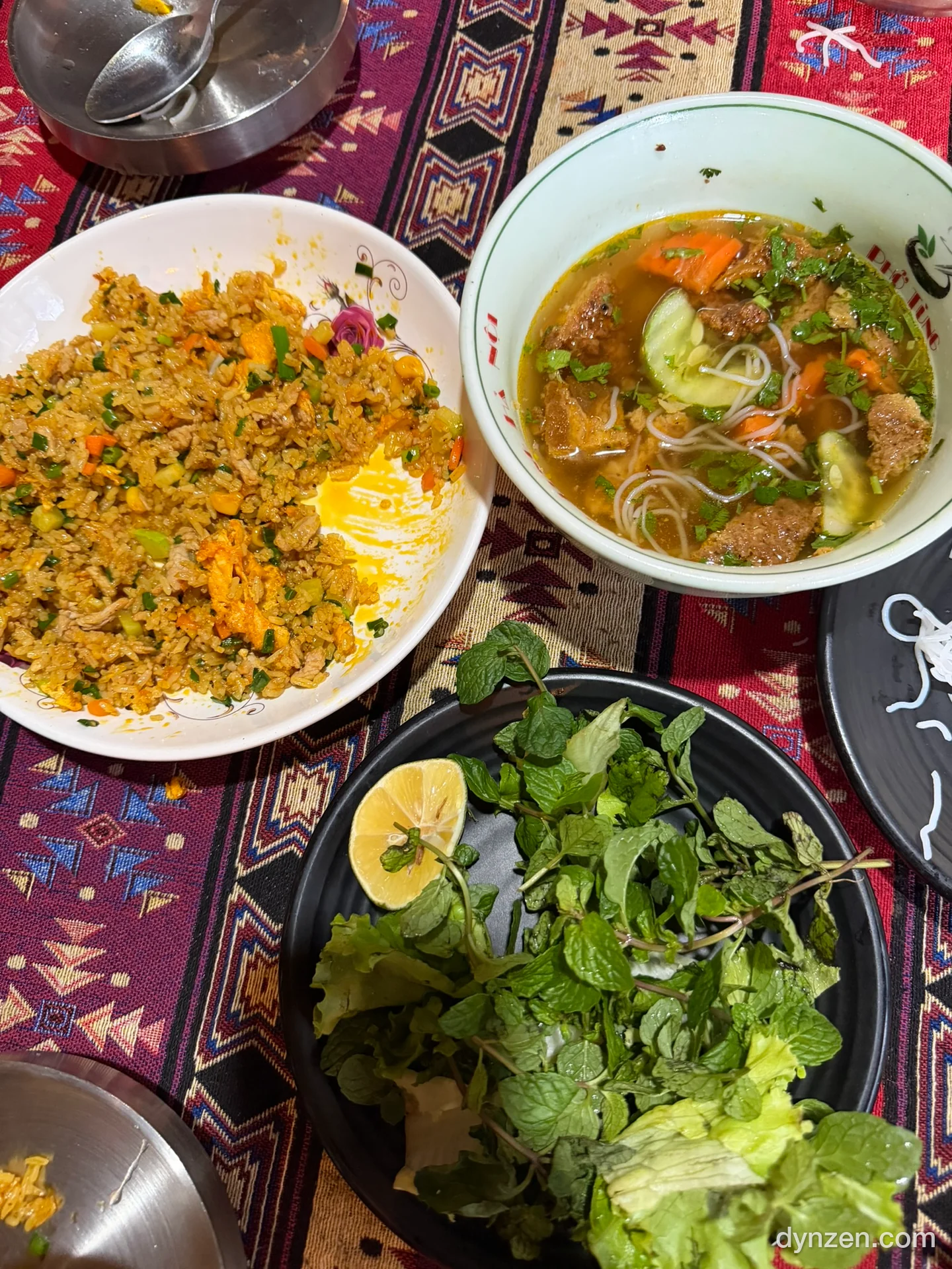 Fried rice and bún chả broth on a table with a H’Mong-pattern tablecloth, rice noodles and a plate of herbs