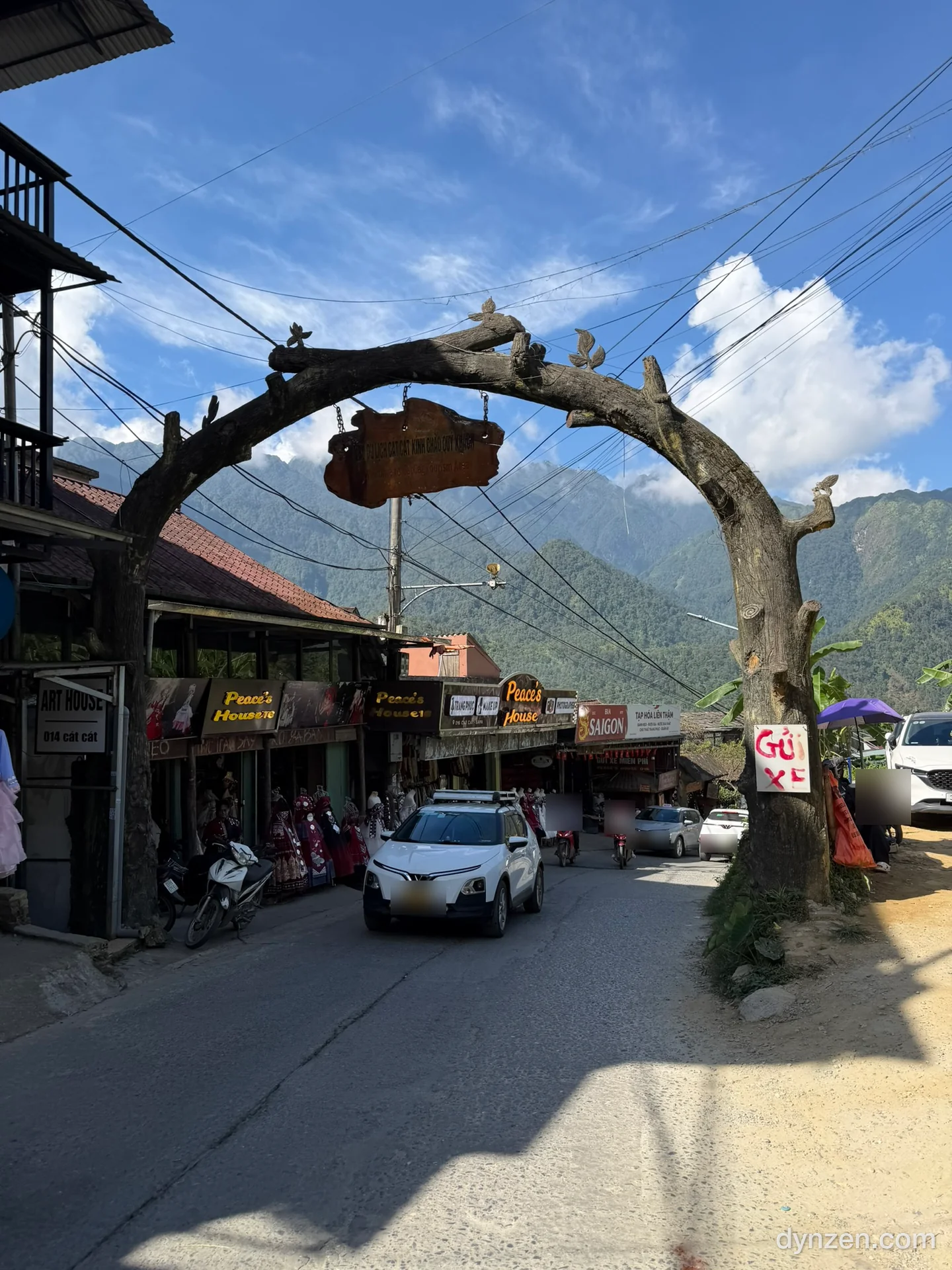 The wooden arch at the entrance of Cat Cat Village, cars and motorbikes passing along a road lined with traditional costume shops
