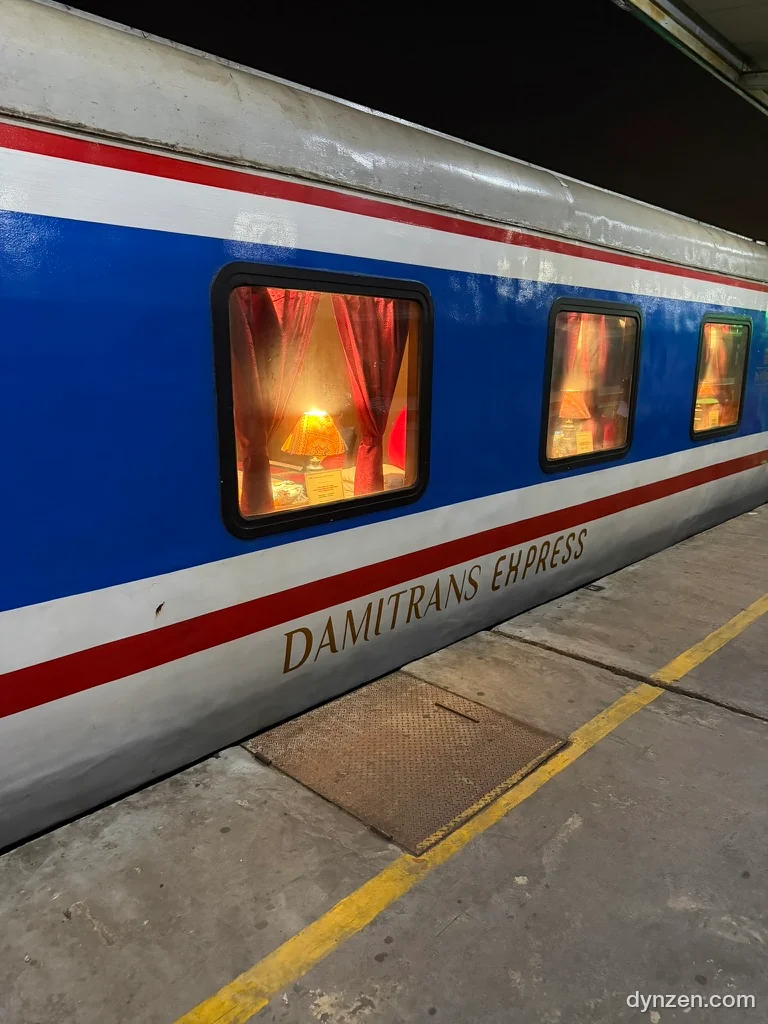 A Damitrans Express carriage at the platform, red curtains and a lamp in the window