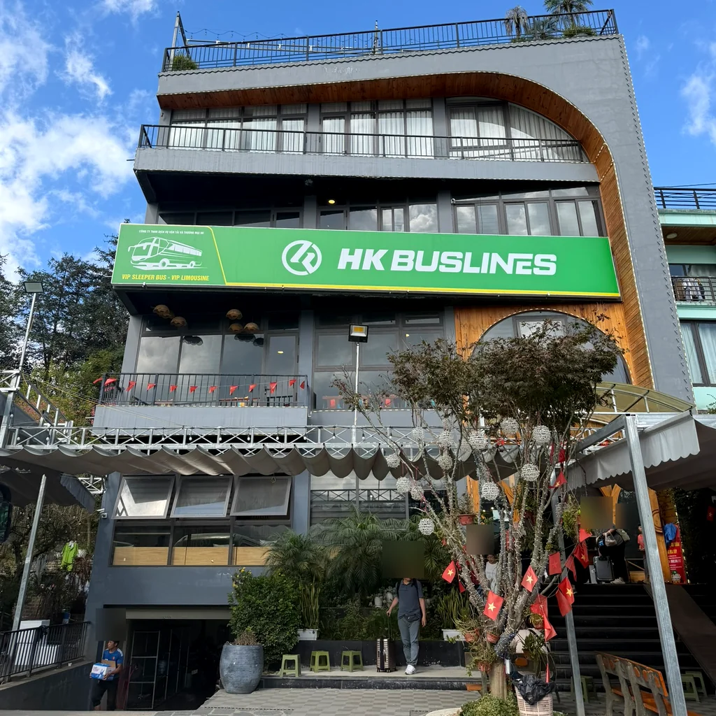 The Sapa office building with a green HK BUSLINES sign under a blue sky, travelers coming and going in front of the steps