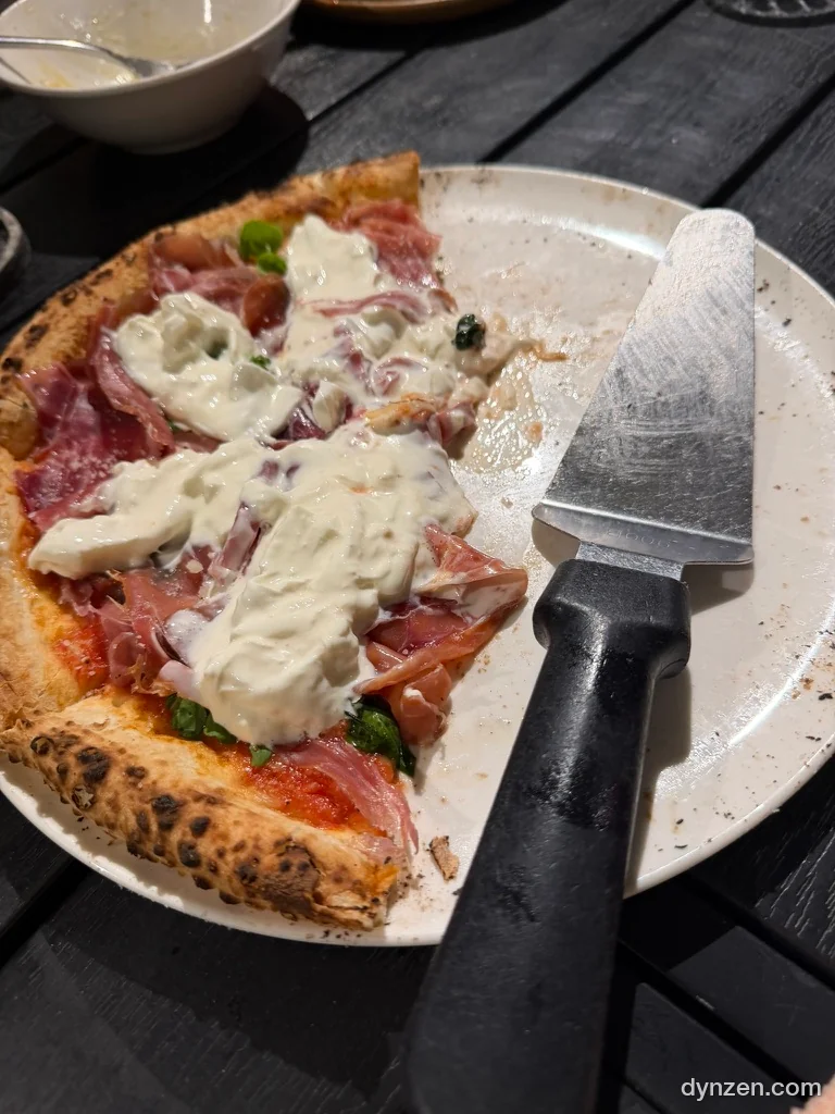 A slice of wood-fired pizza topped with burrata and Parma ham on a white plate, with a pizza server