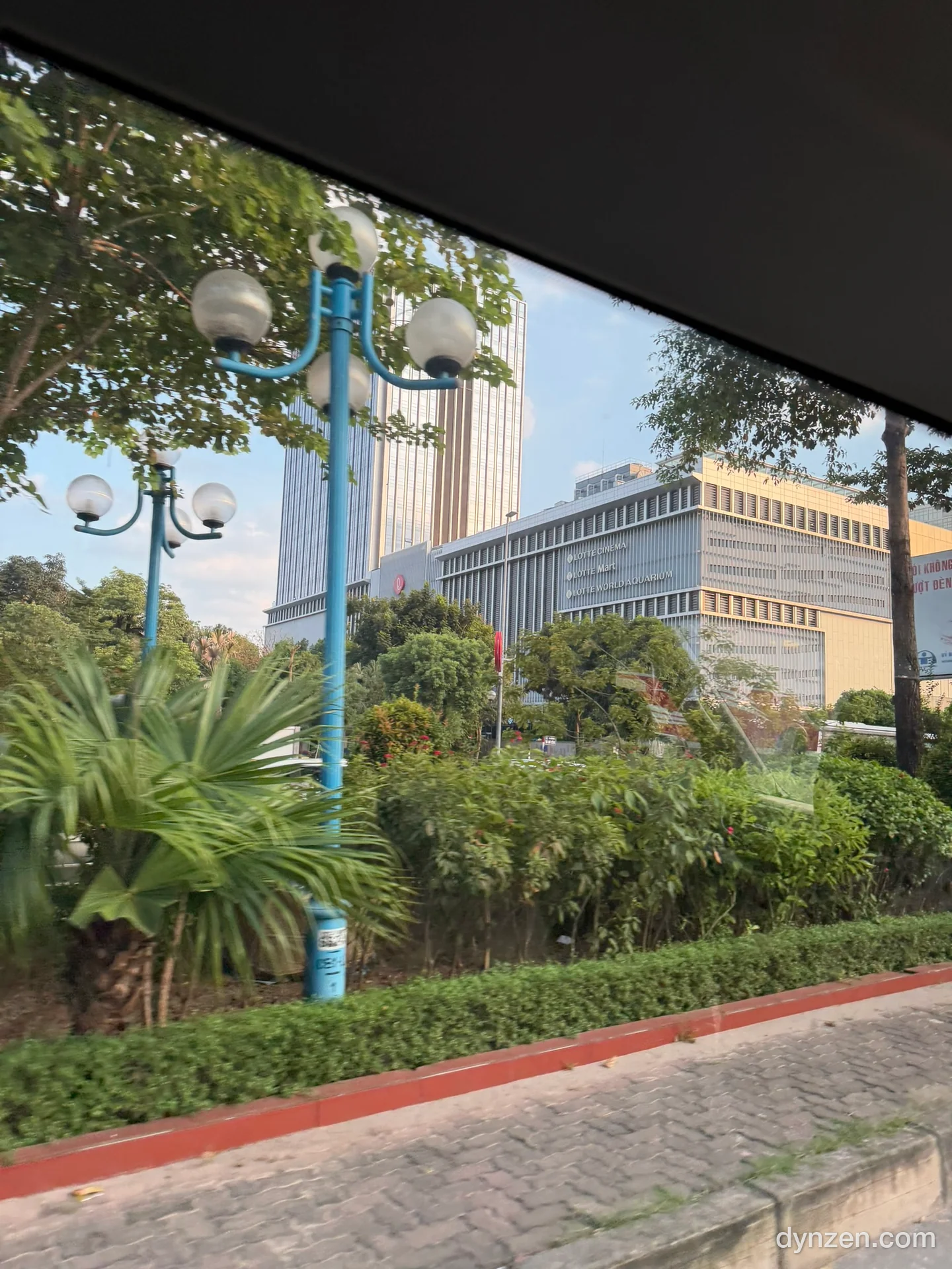 Tall buildings of Lotte Mall West Lake, street trees and a flower bed seen through the shuttle window