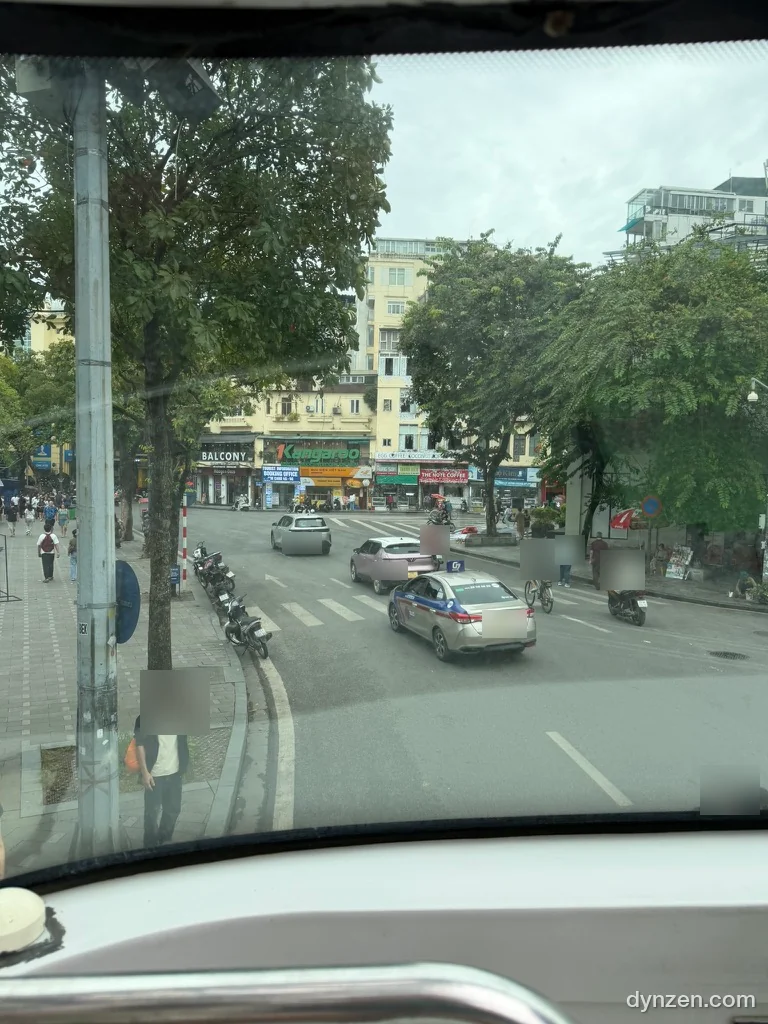 The Hoan Kiem street seen from the front seat of the upper deck, an intersection with taxis and motorbikes and roadside trees