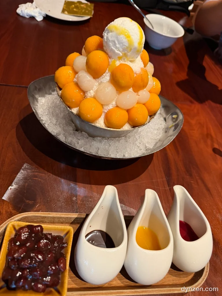 Mango bingsu with round pieces of fresh mango and ice cream piled in a metal bowl, small bowls of red bean and syrup in front