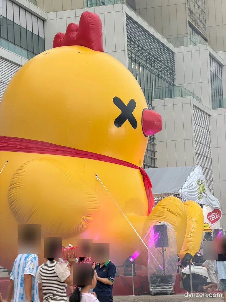 A giant yellow duck balloon in the plaza in front of Lotte Mall West Lake, black X-shaped eyes and a red ribbon at its neck