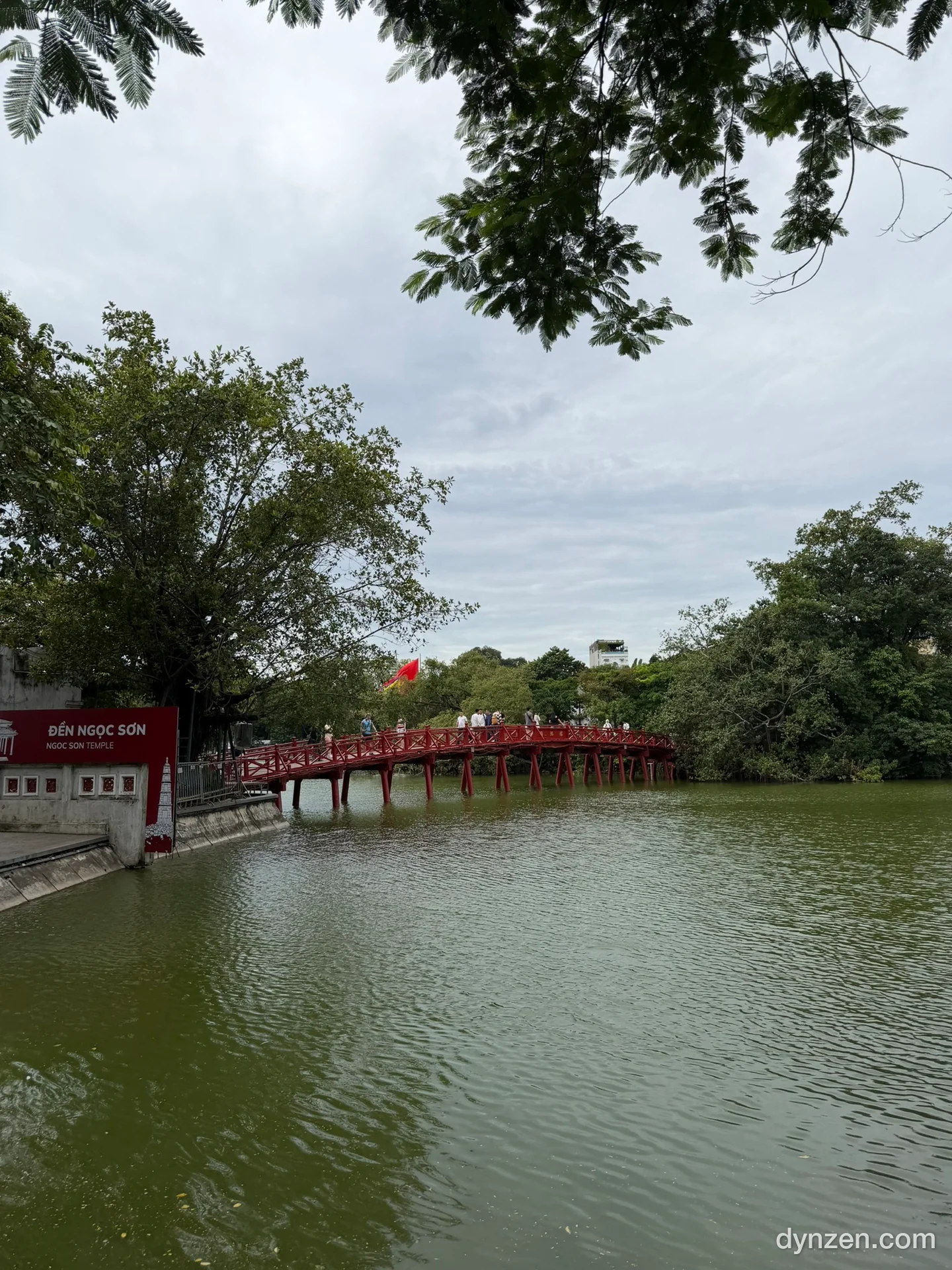 The red The Huc Bridge over Hoan Kiem Lake, trees along the lake edge and a cloudy sky