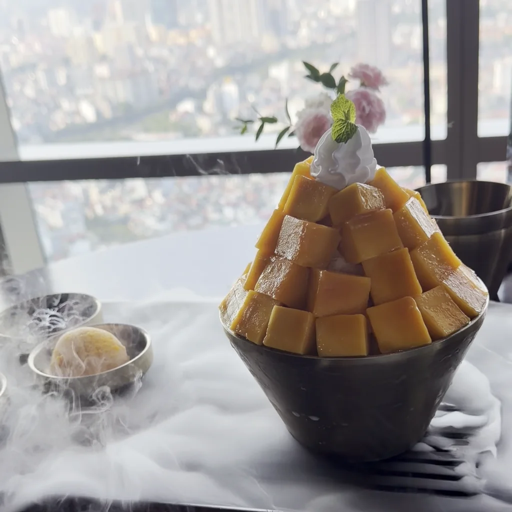 A high-floor window table at Lotte Hotel Hanoi, fresh mango cubes stacked like a pyramid on a bingsu with dry ice smoke spreading below it and the Hanoi skyline outside