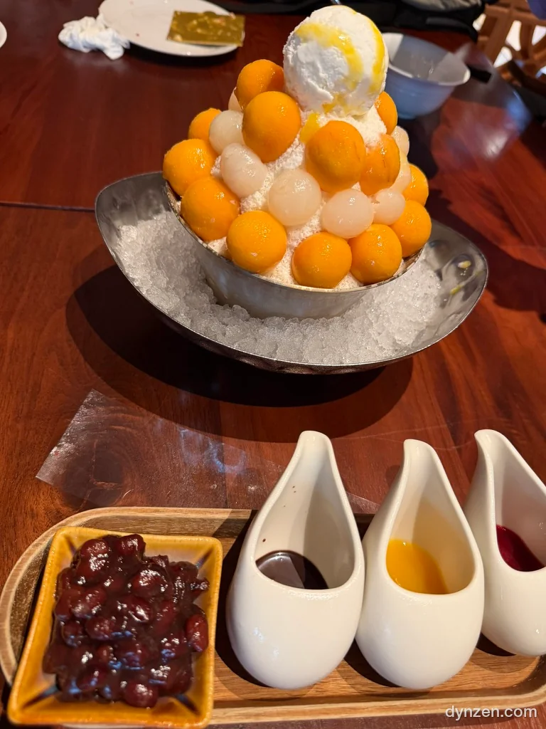 Mango bingsu in a metal bowl, round scoops of fresh mango and lychee piled up with ice cream on top, ice spread around the bowl, red bean and three sauces in front